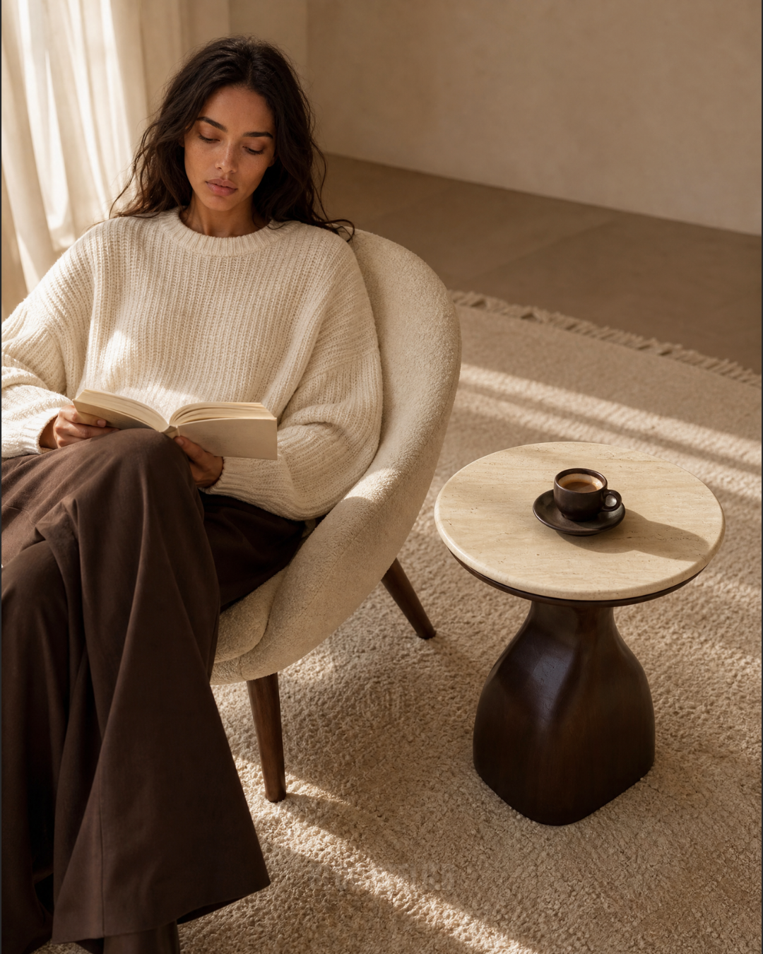 A woman sits in a cream upholstered armchair reading a book in a sunlit room, next to a small round side table with a travertine tabletop with a cup of coffee. She wears a cream ribbed knit sweater and dark brown wide-leg trousers.