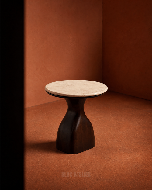 A modern side table with a round light beige travertine top and a sculptural dark brown wooden pedestal base, set against a warm terracotta textured background.