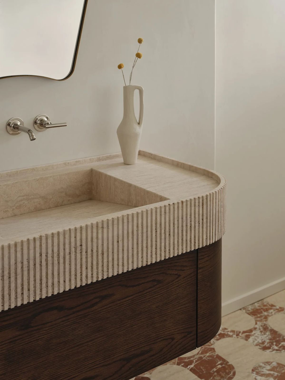 A modern bathroom vanity featuring a curved, ribbed beige travertine stone countertop and sink with a dark wood cabinet base, styled with a white ceramic vase containing dried yellow flowers.
