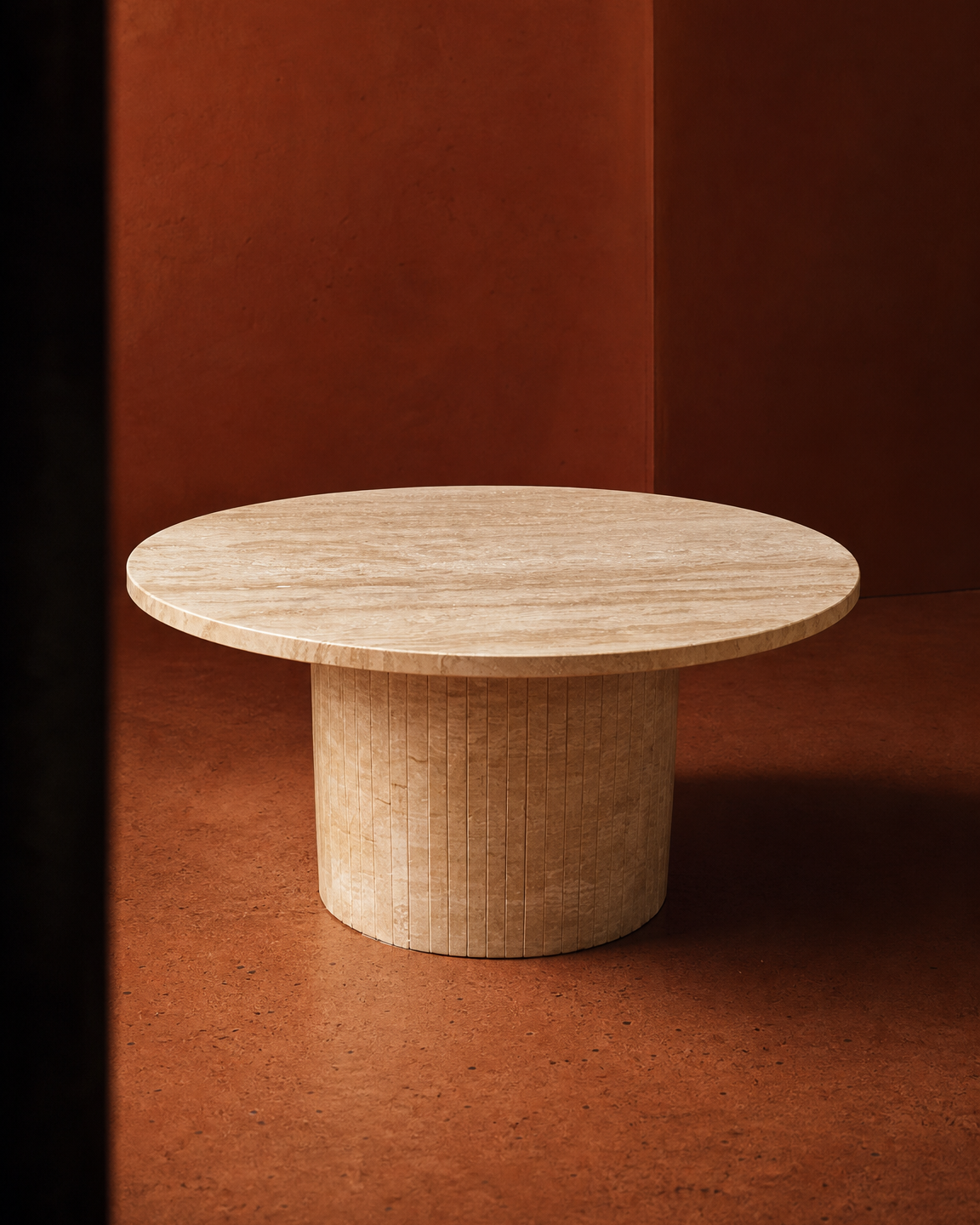A minimalist round travertine coffe table with a thick cylindrical pedestal base, shown in a warm, earthy studio setting with a terracotta-colored background.