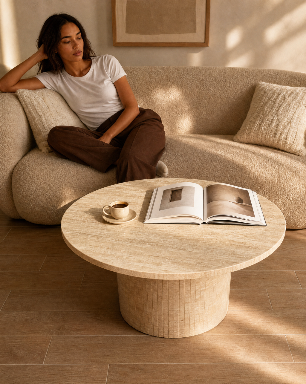A woman relaxes on a textured beige sofa in a sunlit living room, with a round travertine coffee table in front holding an open book and a small cup of coffee.