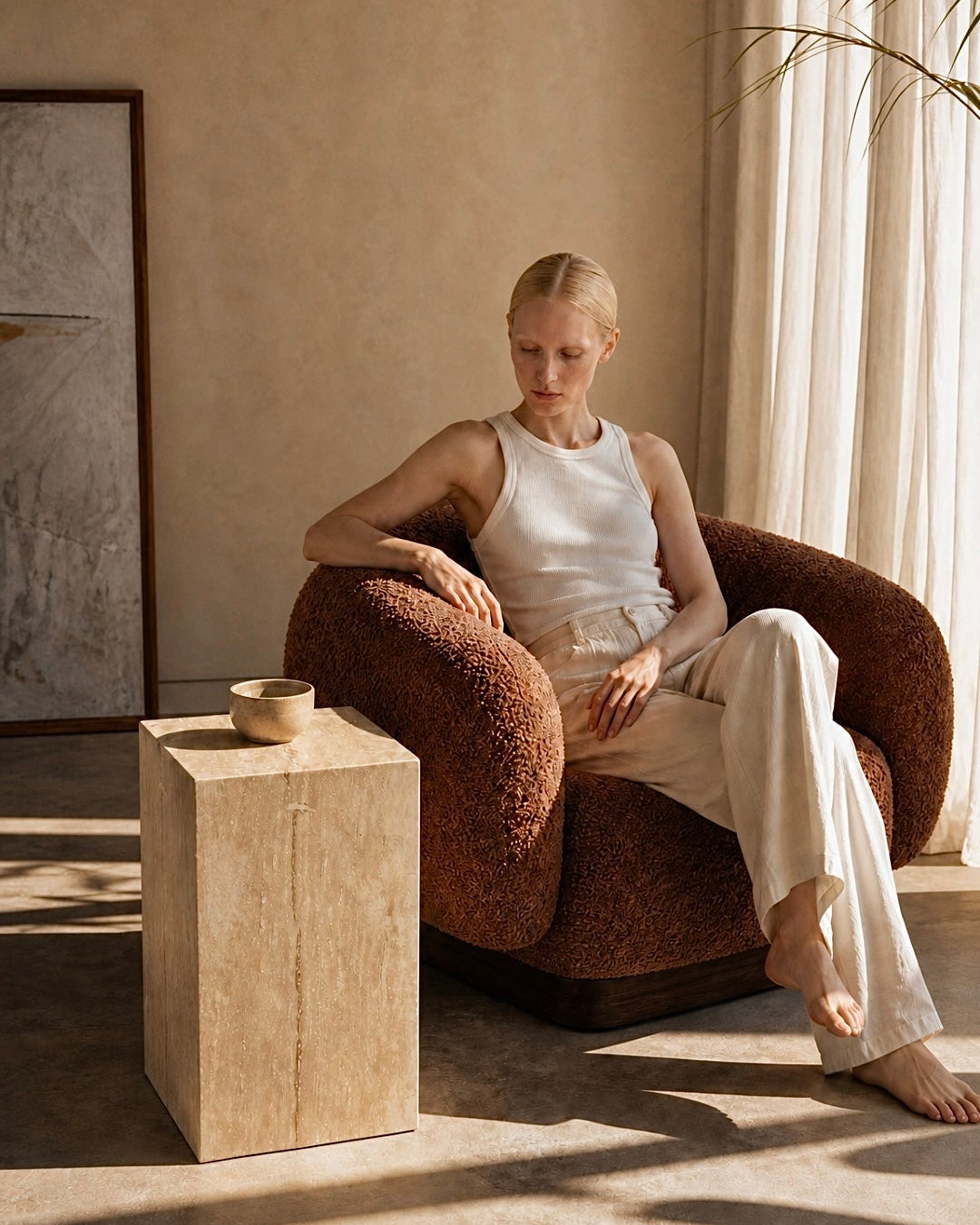 A woman sits barefoot in a textured brown armchair in a sunlit room, wearing a white sleeveless top and white pants. A small travertine stone side table stands beside the chair, and warm natural light streams in from a window with sheer curtains.