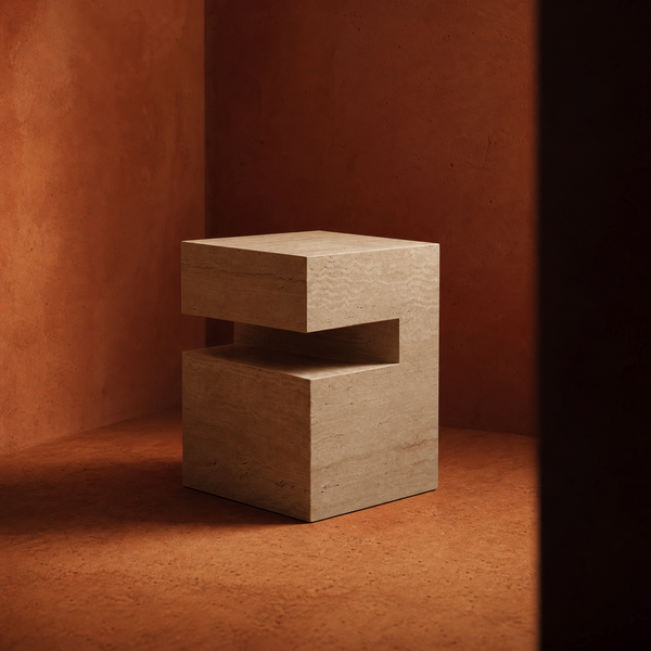 A modern, geometric travertine side table with a stacked block design and a central cutout, set against a warm terracotta textured background.