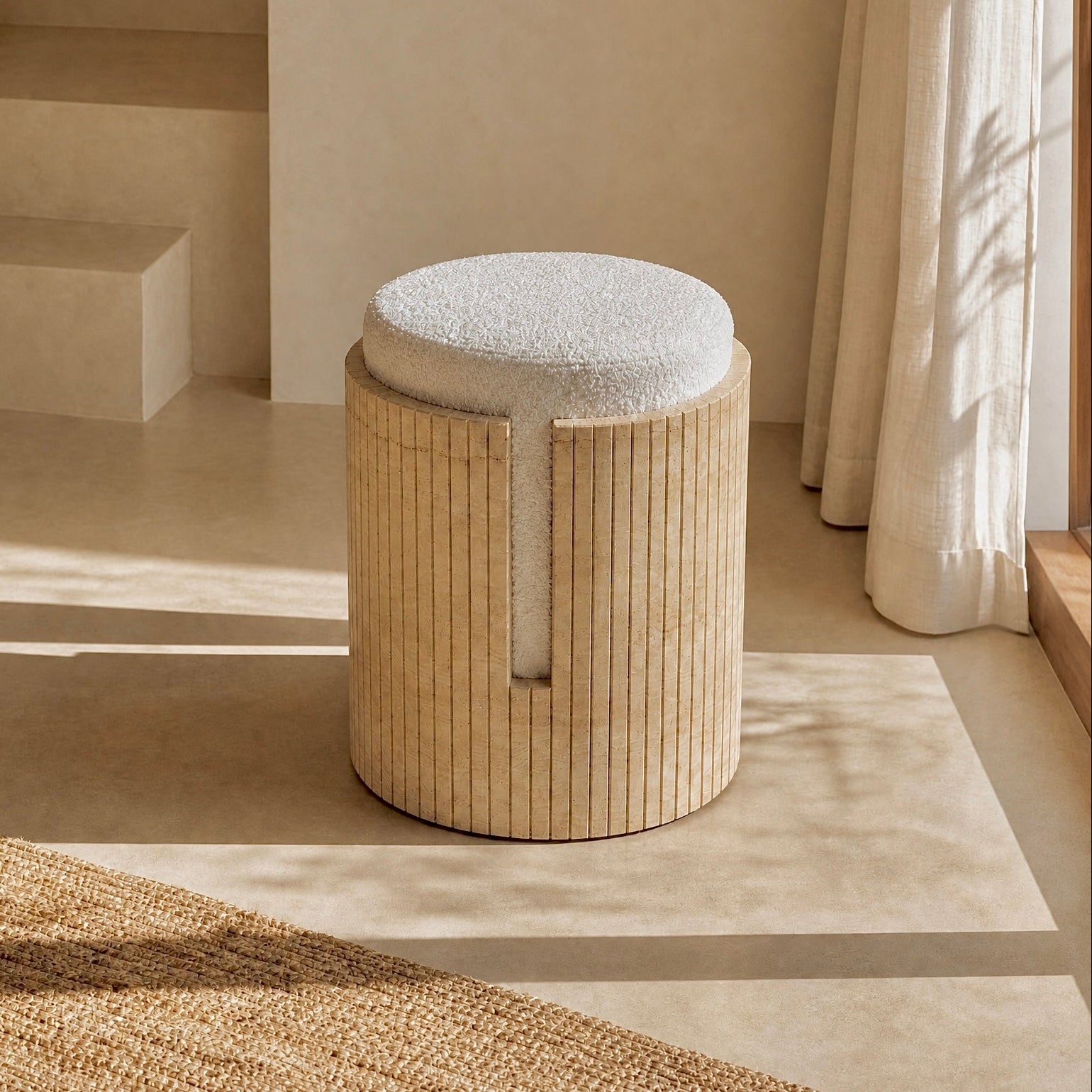 A cylindrical upholstered stool with a light beige travertine fluted frame and a round off-white seat cushion, placed in a softly lit beige interior with steps, curtains, and a woven rug nearby.